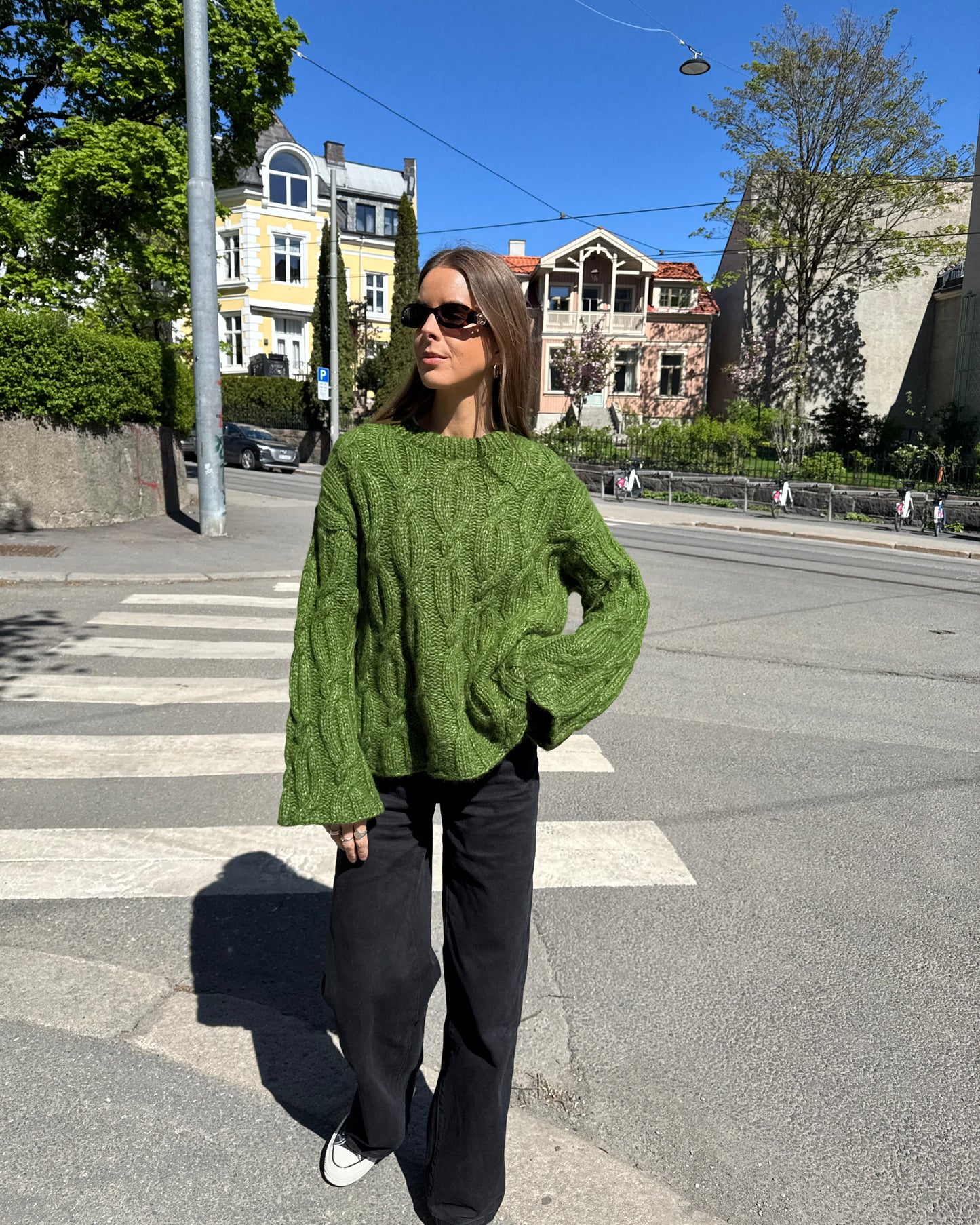Person wearing a green sweater and black pants standing on a street with buildings and trees in the background.