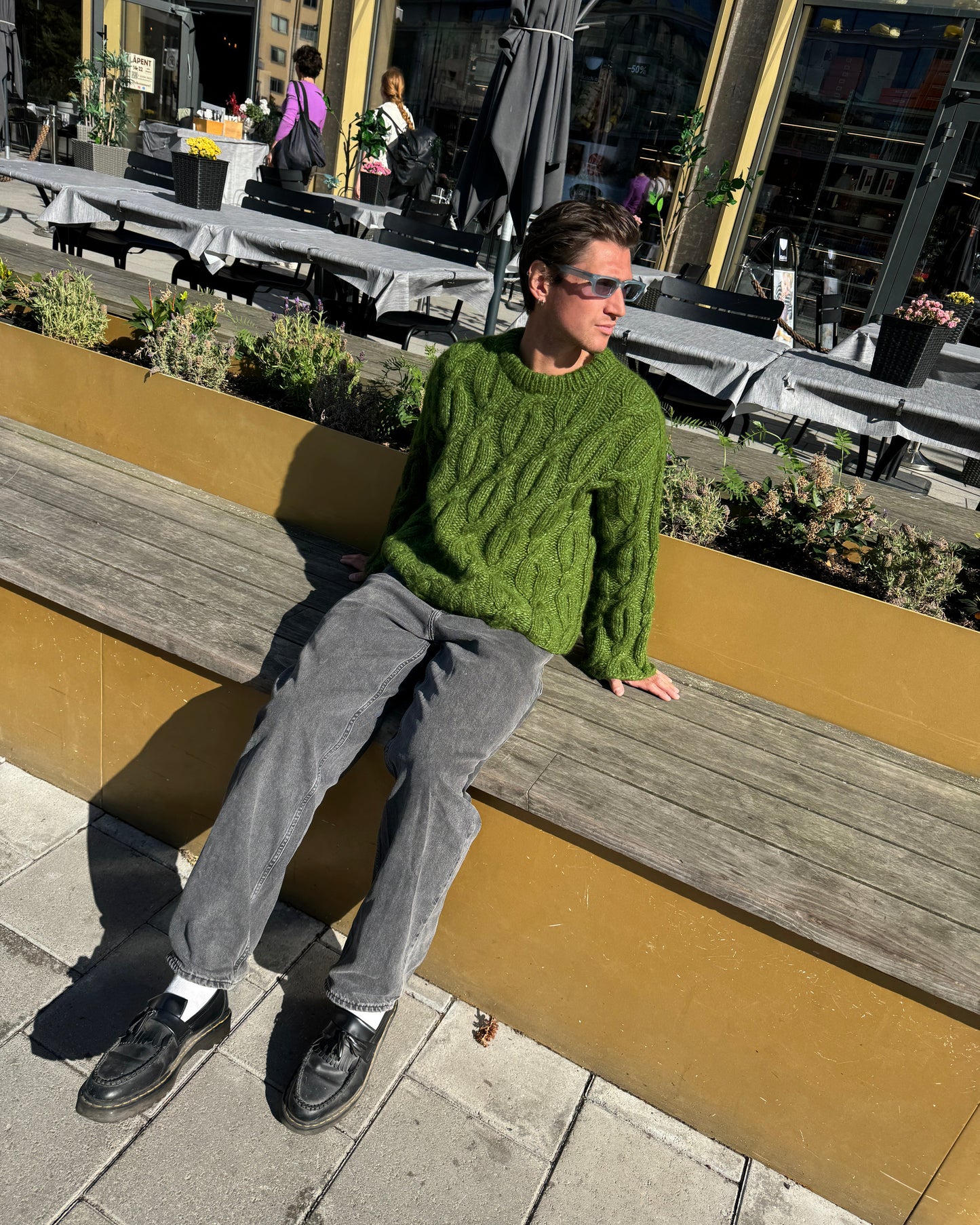 Person in a green sweater and gray pants leaning against a wooden post on a city street.