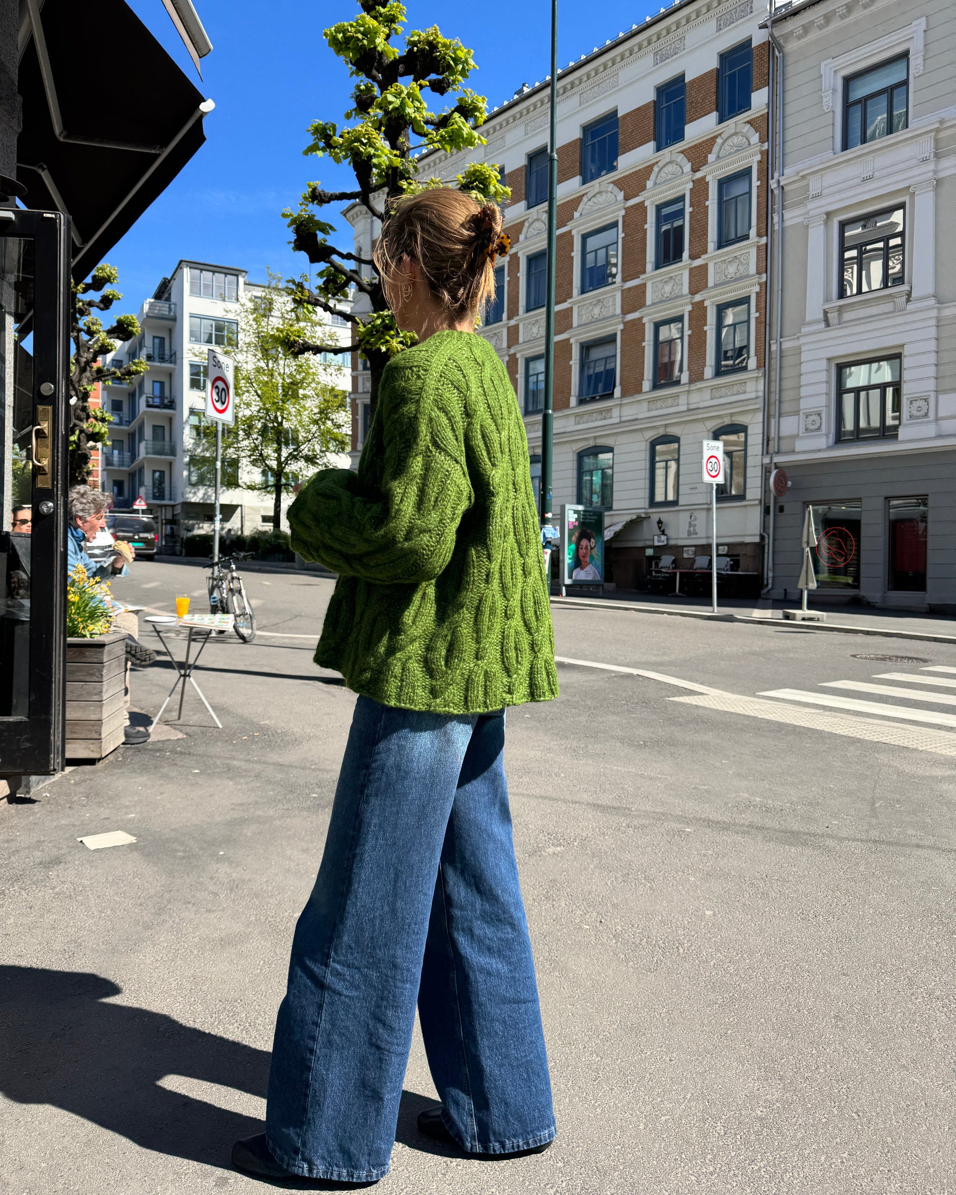 Person wearing a green sweater and blue jeans standing on a city street with buildings in the background.