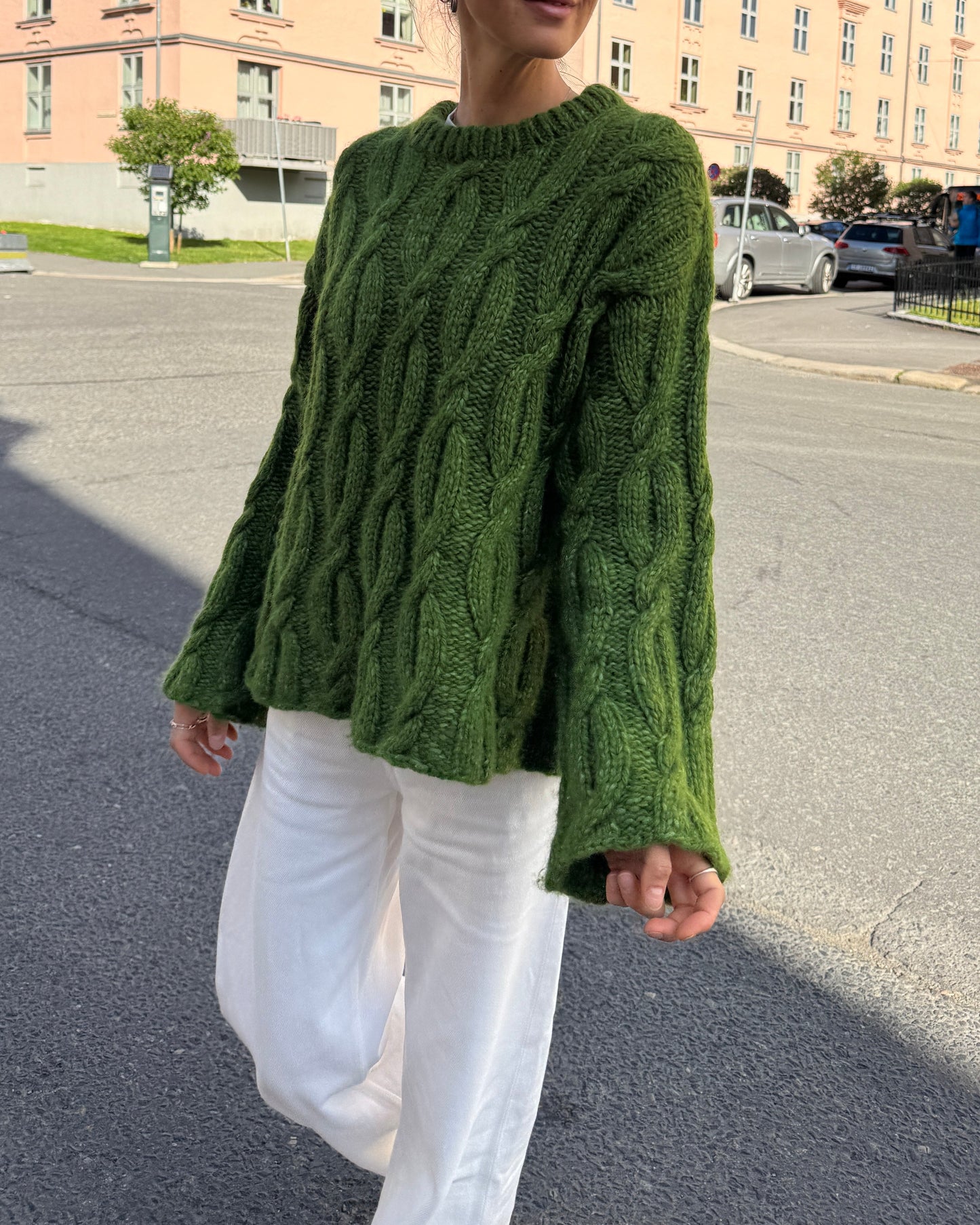 Person wearing a green cable knit sweater and white pants on a street.