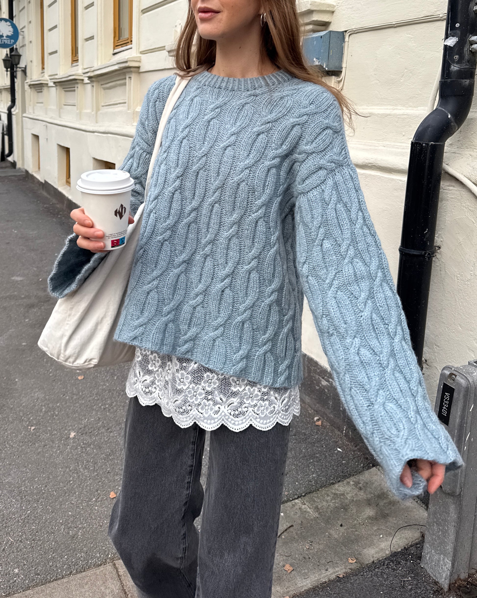 Person wearing a light blue cable knit sweater with a lace top, holding a coffee cup on a street.