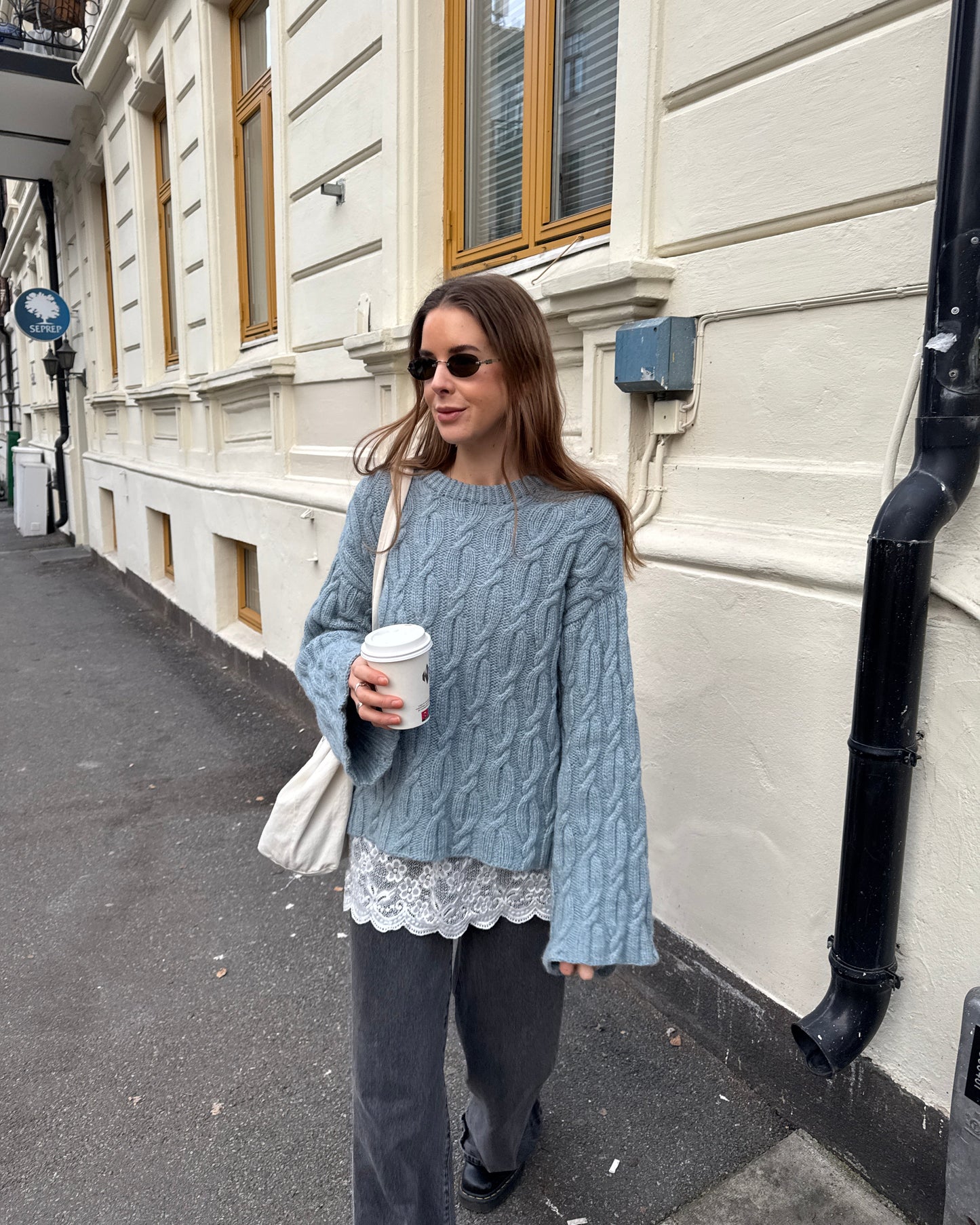 Woman in a gray cable knit sweater with lace trim holding a coffee cup on a street.
