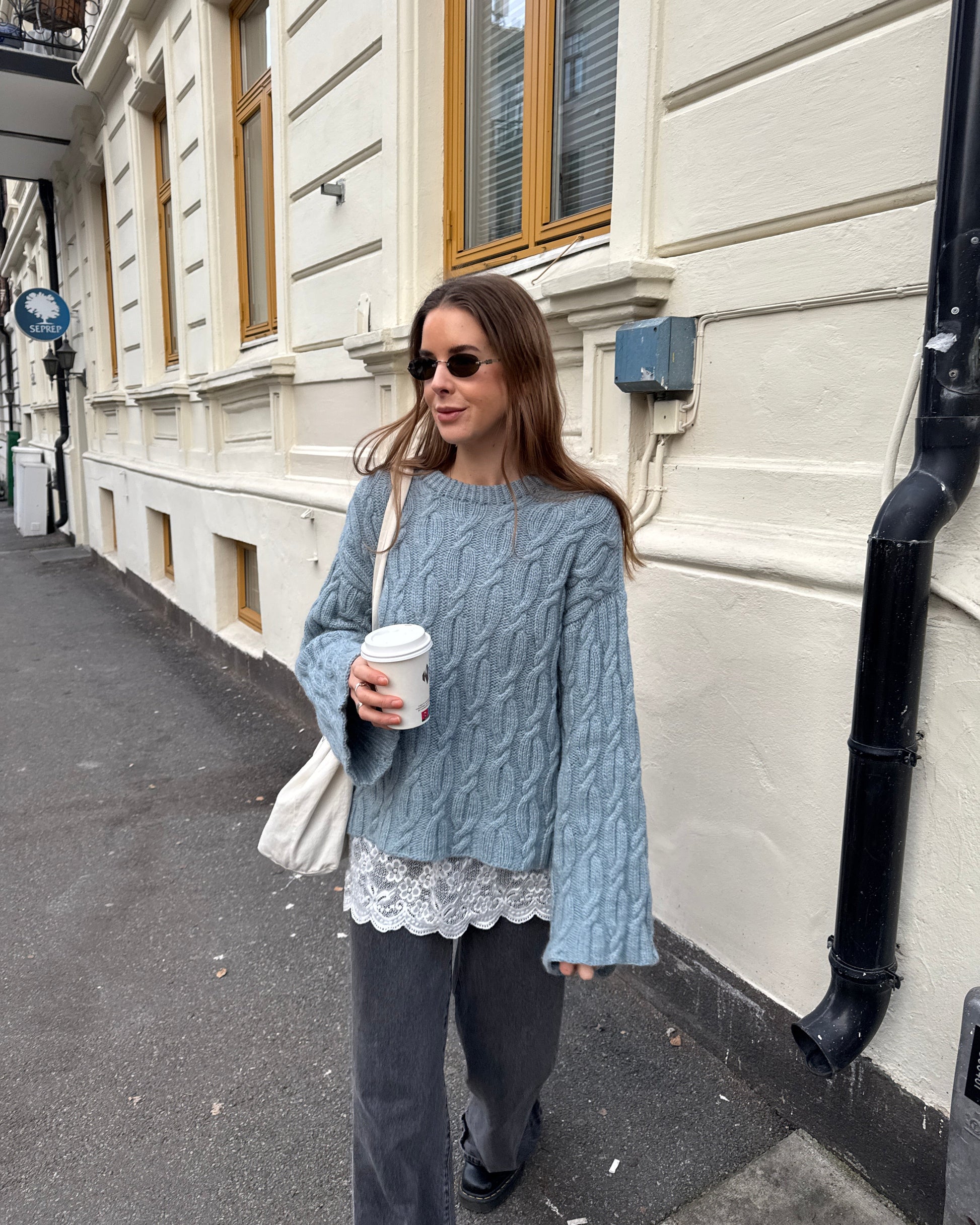 Woman in a gray cable knit sweater with lace trim holding a coffee cup on a street.
