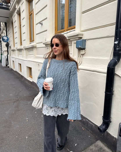 Woman in a gray cable knit sweater with lace trim holding a coffee cup on a street.