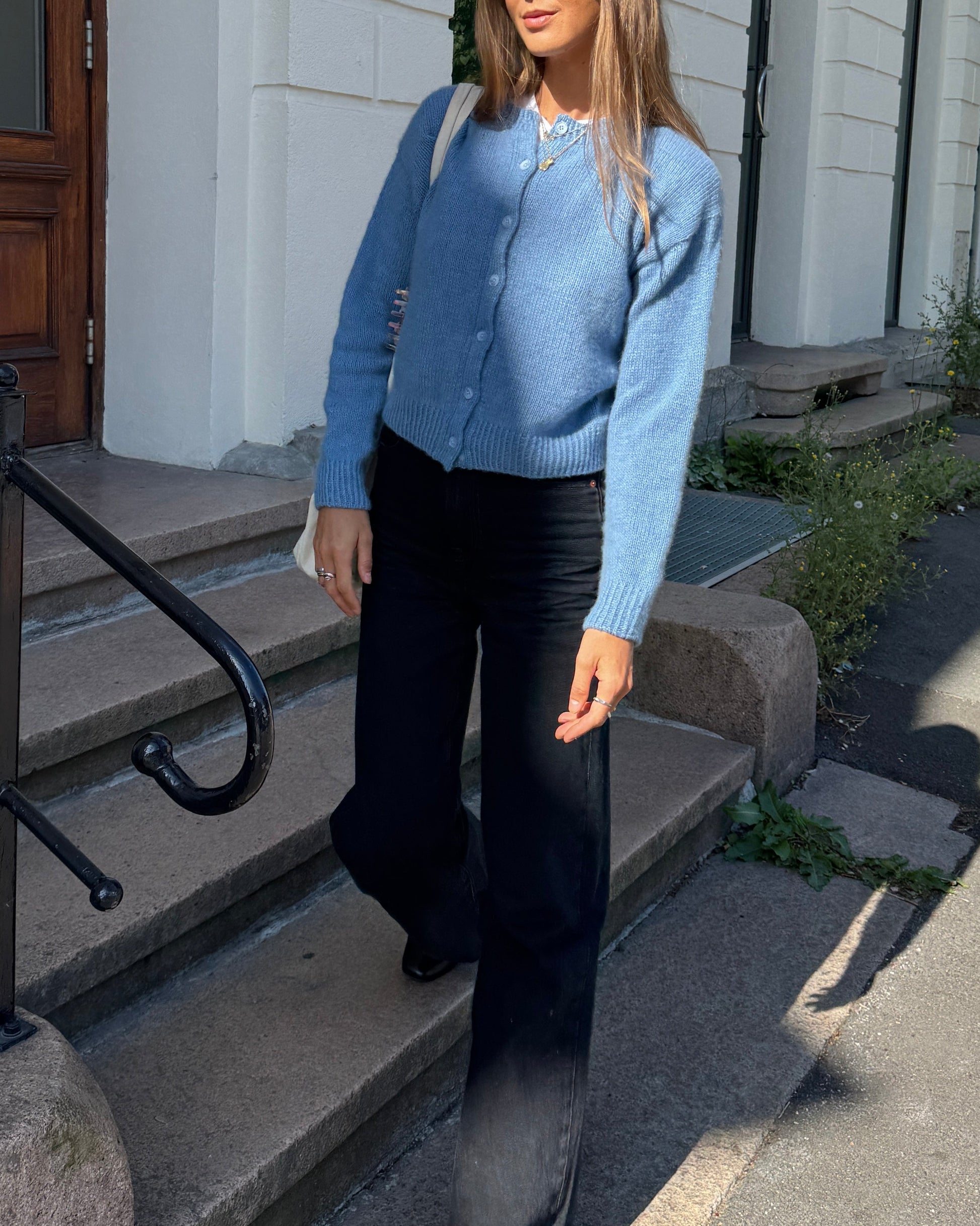 Person wearing a blue cardigan and black pants standing on steps outside a building.